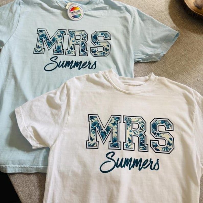 May include: Two short-sleeved t-shirts, one light blue and one white, are displayed. Each shirt features the text "MRS Summers" in a floral design. The light blue shirt has a tag attached. The shirts are likely for a wedding or bridal event.