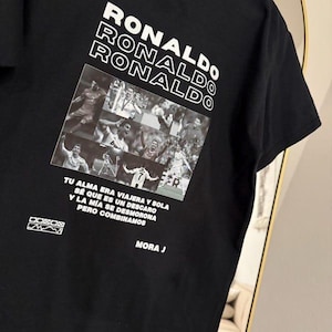 May include: Black t-shirt featuring the name "RONALDO" stacked in white block letters. Below, a collage of images of a soccer player. The shirt includes Spanish text and the name "MORA J".