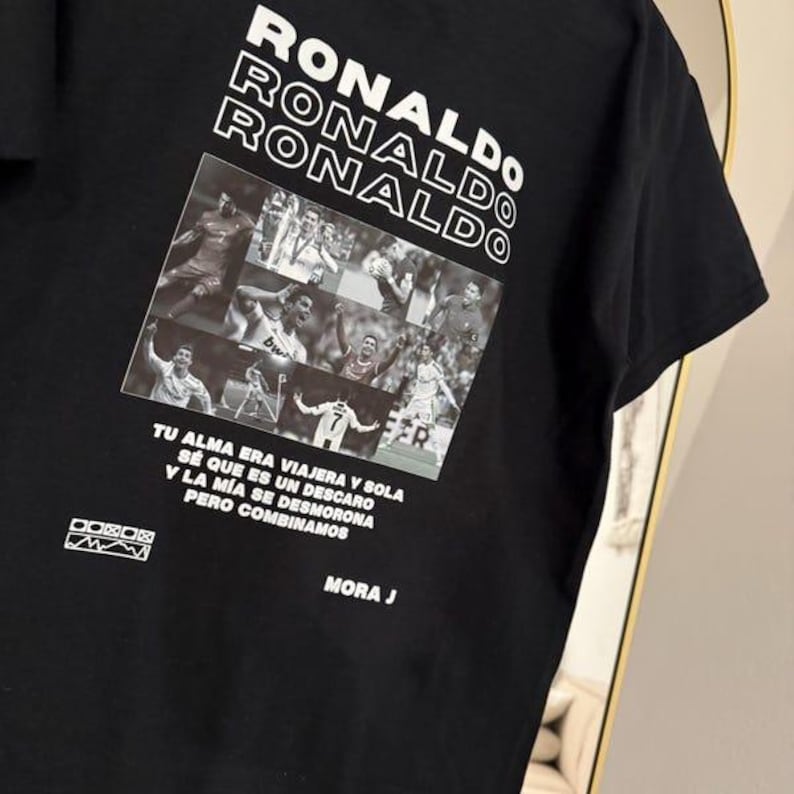 May include: Black t-shirt featuring the name "RONALDO" stacked in white block letters. Below, a collage of images of a soccer player. The shirt includes Spanish text and the name "MORA J".