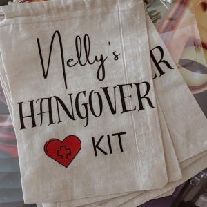 May include: Off-white canvas bags with black script that reads "Nelly's HANGOVER KIT". A red heart with a white cross is below the text. The bags are stacked on top of each other.
