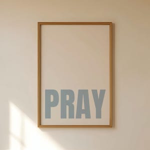 May include: A rectangular wall art print in a light wood frame featuring a cream-coloured background with the word PRAY printed in large, bold, slate-blue capital letters centred in the lower portion of the vertical composition.