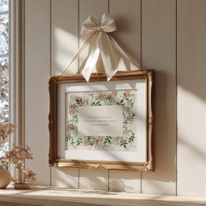 May include: A rectangular gold-toned ornate frame containing a print with a floral border of green leaves and peach flowers. The center text reads: My people will live in peaceful dwelling places, in secure homes. Isaiah 32:18. A cream satin ribbon bow hangs above.