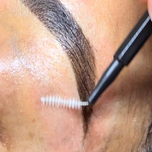 Pode incluir: Uma ferramenta preta para cuidar das sobrancelhas com uma pequena ponta de escova branca, usada para pentear e modelar os pelos das sobrancelhas castanho-escuras. A ferramenta possui um cabo preto fino e uma cabeça de cerdas brancas finas.