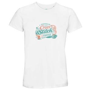 May include: White t-shirt featuring the text "Powered by Cross Stitch & Coffee" in a colourful design. The graphic includes a thread spool, needle, and a coffee cup. The shirt is made of a soft material.