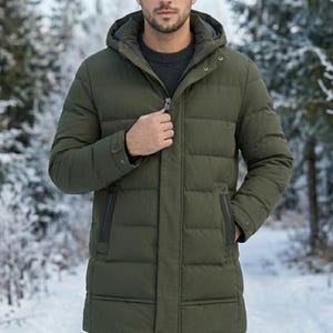 May include: An olive green winter coat with a hood and a zip. The coat has a quilted design and pockets. The person in the image is wearing a dark grey sweater. The background is a snowy forest.
