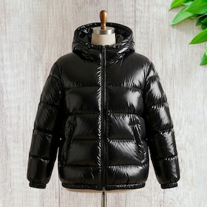 Puede incluir: Una chaqueta acolchada negra brillante con capucha, exhibida en un maniquí. La chaqueta tiene una cremallera completa, dos bolsillos laterales y puños acanalados. El material tiene una calidad brillante y reflectante.