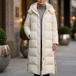 May include: A long, cream-coloured puffer coat with a hood. The coat features a quilted design, a zip closure, and side slits. The person is wearing a grey turtleneck sweater underneath. The coat appears to be made of a lightweight, water-resistant material.