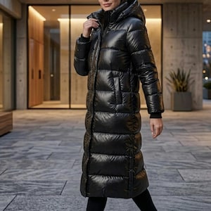 May include: A long, black puffer coat with a hood. The coat is floor-length and has a quilted design. It features a zip closure and pockets. The model is wearing black trousers and a black top. The coat appears to be made of a shiny, water-resistant material.