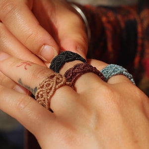 Puede incluir: Primer plano de cuatro anillos de macramé hechos a mano en varios colores: beige, marrón oscuro, negro y verde azulado. Los anillos están en los dedos de una persona, con otra mano ajustando uno. Los anillos tienen una textura tejida.