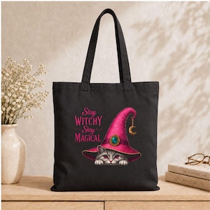 May include: A black tote bag featuring a graphic of a gray cat wearing a bright pink witch hat with a gold crescent moon charm and a teal gemstone. The text "Stay WITCHY Stay MAGICAL" is printed in pink above the cat illustration.