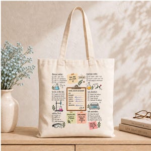 Op de afbeelding: Een crèmekleurige canvas draagtas met een kleurrijke grafische print van een laboratoriumnotitieboekpagina voor organische chemie. Het ontwerp bevat diverse wetenschappelijke iconen, checklists en handgeschreven notities over labprocedures.