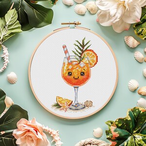 May include: A circular wooden embroidery hoop containing a cross-stitch pattern of an orange cocktail in a stemmed glass. The drink features a striped straw, a sprig of greenery, and an orange slice garnish, with a smiling face on the glass.