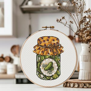 Op de afbeelding: Een ronde houten borduurring met een kruissteekpatroon van een glazen pot gevuld met groene augurken. Het deksel is geel met een klein bloemmotief en een gestrikt lint. Het etiket in het midden toont een augurk en een takje dille.