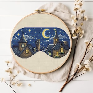 May include: A circular wooden embroidery hoop containing a cross-stitch pattern of a night scene with houses, a cat sitting on a chimney, a crescent moon, and stars against a dark blue sky. The fabric is light beige and the hoop has a metal screw.