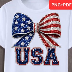 May include: A white t-shirt featuring a large graphic of a bow decorated with a red, white, and blue American flag pattern made of sparkling rhinestones. Below the bow, the letters USA are printed in a bold, blue, rhinestone-encrusted font.