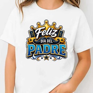 May include: White t-shirt featuring a colorful graphic design. The design includes the words "Feliz Dia Del Padre" in blue and gold, a mustache, and a crown. The shirt is a casual wear item.