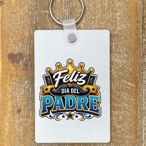 May include: A white rectangular keychain with a silver keyring. The keychain features a colorful graphic with the words "Feliz Dia Del Padre" in blue and gold, with a crown and mustache design.
