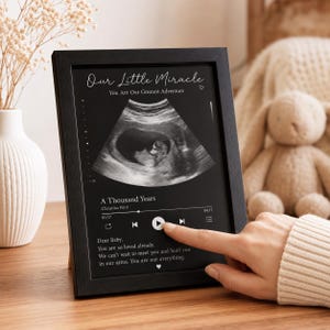 Puede incluir: Un marco rectangular negro que contiene una impresión personalizada con una imagen de ecografía, el texto Our Little Miracle, You Are Our Greatest Adventure, A Thousand Years de Christina Perri y un mensaje cariñoso para un bebé.