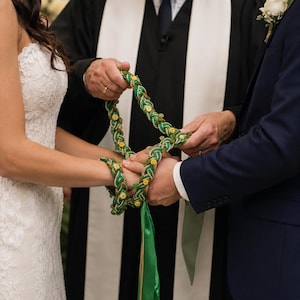 Puede incluir: Ceremonia de boda con una cuerda de handfasting verde y dorada. La novia lleva un vestido de encaje blanco y el novio un traje azul marino. Un religioso con una túnica negra y blanca oficia la ceremonia.