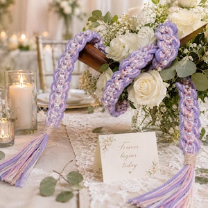 Puede incluir: Primer plano de un ramo de novia con rosas blancas y vegetación. Dos piezas de macramé lavanda con detalles de perlas adornan el ramo. Una tarjeta con el texto "Forever begins today" está sobre la mesa.
