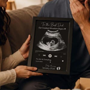 Könnte beinhalten: Ein schwarz gerahmter Druck mit einem Ultraschallbild in Graustufen, dem Text To the Best Dad, Our Greatest Adventure Begins, einer Musik-Player-Oberfläche mit dem Titel You Are My Everything, Gummy Bear und Baby Arriving January 2026.