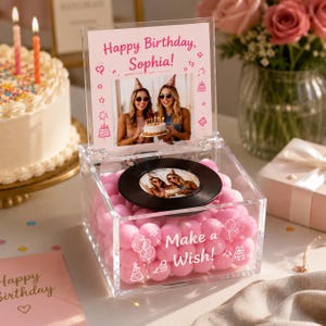 Könnte beinhalten: Eine quadratische Box aus klarem Acryl, gefüllt mit rosa Pompons. Der Deckel zeigt ein Foto von zwei Personen und den Text Happy Birthday, Sophia!. Oben liegt ein schwarzer Vinyl-Schallplatten-Einsatz, vorne steht der Text Make a Wish.