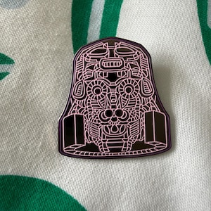 Peut inclure: Une broche en émail métallique avec une base violet foncé et des lignes rose clair complexes formant un visage stylisé et symétrique rappelant un masque. Le motif présente des formes géométriques, des yeux circulaires et des éléments latéraux.