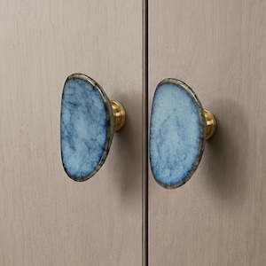 May include: Two cabinet knobs with a blue, agate-like design and brass hardware. The knobs are mounted on a light-colored wooden cabinet door. The knobs have a unique, organic shape, adding a touch of elegance.