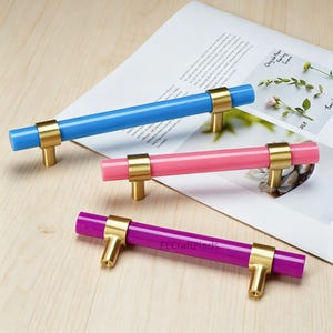 May include: Three cabinet pulls in blue, pink, and purple. Each pull has a cylindrical colored center and gold-toned metal hardware. The pulls are arranged on a light-colored wooden surface.