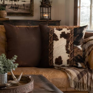 Two square throw pillows. One is solid dark brown with a textured surface. The other features a dark brown side, a central panel of white and brown cowhide print, and a right side of brown and black cowhide print , joined by realistic looking printed laced leather strips.