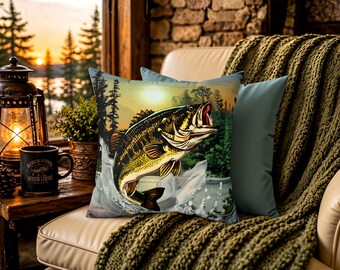 May include: A square throw pillow featuring a graphic illustration of a largemouth bass jumping out of water, set against a backdrop of pine trees and a sunset sky. The pillow is positioned on a light-colored chair with a green knit blanket.