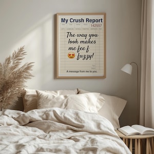 May include: A rectangular wall art print titled My Crush Report with a light beige background and black grid lines. The text reads The way you look makes me feel fuzzy with a heart-eyes emoji. It is framed in a light wood frame.