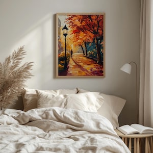 May include: A framed art print depicts a park scene in autumn. The artwork features a pathway with a person walking towards a lamppost. Trees with orange and red leaves line the path. The artwork is displayed above a bed with beige bedding.