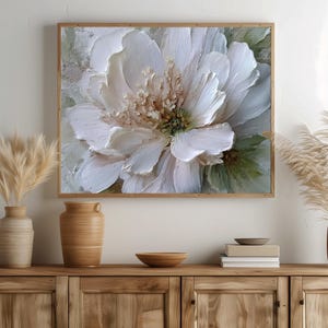 Op de afbeelding: Een ingelijst, getextureerd schilderij van een grote witte bloem met gedetailleerde bloemblaadjes en een getextureerd centrum. Het kunstwerk heeft een lichtgroene en beige achtergrond en is gevat in een natuurlijke houten lijst. De schildering is een close-up van de bloem.