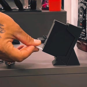Puede incluir: Un soporte negro, rectangular e inclinado sostiene un pequeño dispositivo electrónico cuadrado y oscuro. Una tira rectangular blanca y delgada se está insertando en el lateral del dispositivo. El soporte tiene una base geométrica sólida.