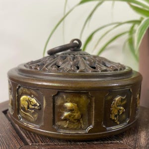 Pode incluir: Um queimador de incenso redondo de bronze com uma tampa decorativa vazada e uma alça de anel central. O corpo cilíndrico é adornado com uma série de painéis em relevo que retratam vários animais do zodíaco chinês em acabamento metálico.