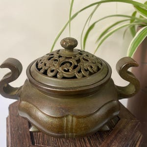 May include: A bronze-toned metal incense burner featuring a rounded body with two curved side handles and a removable lid. The lid displays an intricate openwork scroll pattern and a small central knob. The base has three small feet.