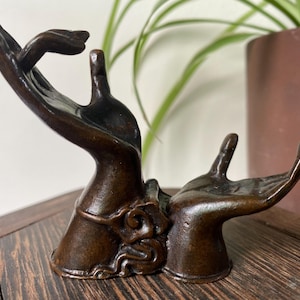 Puede incluir: Una escultura de color bronce oscuro que presenta dos manos posicionadas juntas. Una mano está levantada con los dedos extendidos hacia arriba, mientras que la otra mano está colocada horizontalmente con la palma hacia arriba. La base presenta detalles decorativos en espiral.
