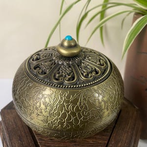 Pode incluir: Um queimador de incenso redondo em metal cor de latão antigo, com um intrincado padrão floral gravado no corpo e uma tampa decorativa perfurada com uma pequena pedra de cor turquesa no botão superior.