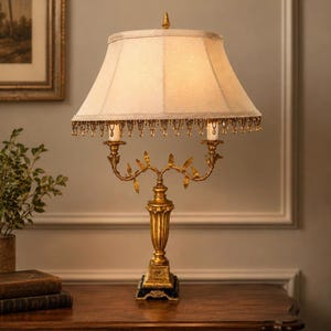 Può includere: Lampada da tavolo decorativa con base dorata e paralume color crema. La lampada presenta un design ornato e dettagliato con un bordo di perline sul paralume. È posizionata su una superficie di legno, alta circa 60 cm.