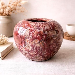 May include: A round, red and white marbled ceramic vase with a wide opening. The vase has a glossy finish and sits on a white surface. Other decorative items are in the background.
