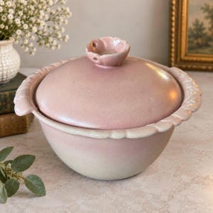 May include: A round ceramic lidded bowl in a soft dusty pink hue. The bowl features a scalloped rim and two side handles. The lid is topped with a small, sculpted flower-shaped knob. The glaze transitions from a deeper pink at the top to a lighter, pale tone at the base.