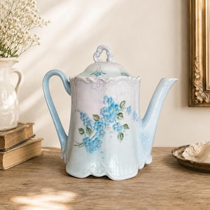 May include: A porcelain teapot with a pale blue and white gradient finish, featuring a hand-painted floral pattern of small blue flowers with green leaves. The teapot has a scalloped base, a curved handle, a tapered spout, and a matching lid.
