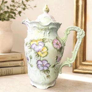 May include: A pale green porcelain pitcher with a lid, featuring embossed scrollwork details and hand-painted purple and yellow pansy flowers. The pitcher has a curved handle, a scalloped rim, and a spout, showcasing a delicate floral design.