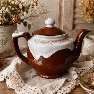 May include: A brown and white ceramic teapot with a lid and handle. The teapot is sitting on a lace doily. Dried flowers and a small teacup are in the background.
