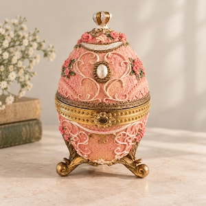 May include: A pink egg-shaped trinket box with a textured surface, adorned with small pink rose accents, white scrollwork, and a central oval pearl-like detail. The container features a gold-toned metal rim and stands on three ornate gold-toned feet.