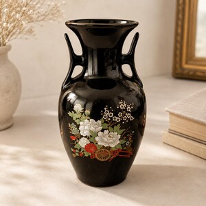 Puede incluir: Un jarrón de cerámica negro brillante con dos asas laterales y un borde acampanado. La superficie frontal está decorada con un motivo floral detallado en blanco, rojo y verde, que incluye peonías y el diseño de una pequeña rueda de carro.