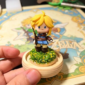 May include: A figurine of a character with blonde hair, blue eyes, and a blue and brown outfit, holding a small branch with green leaves. The figurine stands on a wooden base covered in green and yellow foliage.