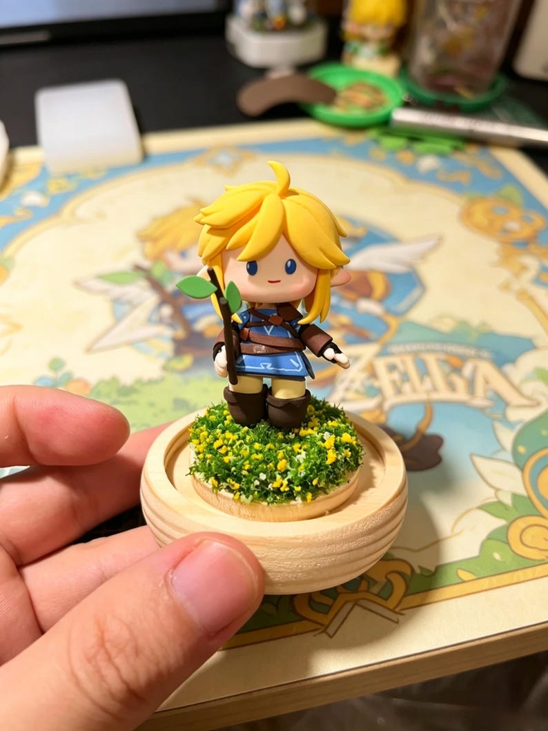 May include: A figurine of a character with blonde hair, blue eyes, and a blue and brown outfit, holding a small branch with green leaves. The figurine stands on a wooden base covered in green and yellow foliage.