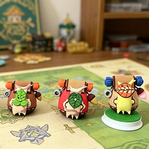 May include: Four colorful, whimsical figurines resembling backpacks with faces, set on a game board. Each figure has unique features, including leaves, orange accents, and different colored bodies. The background includes art supplies and a wooden shelf.